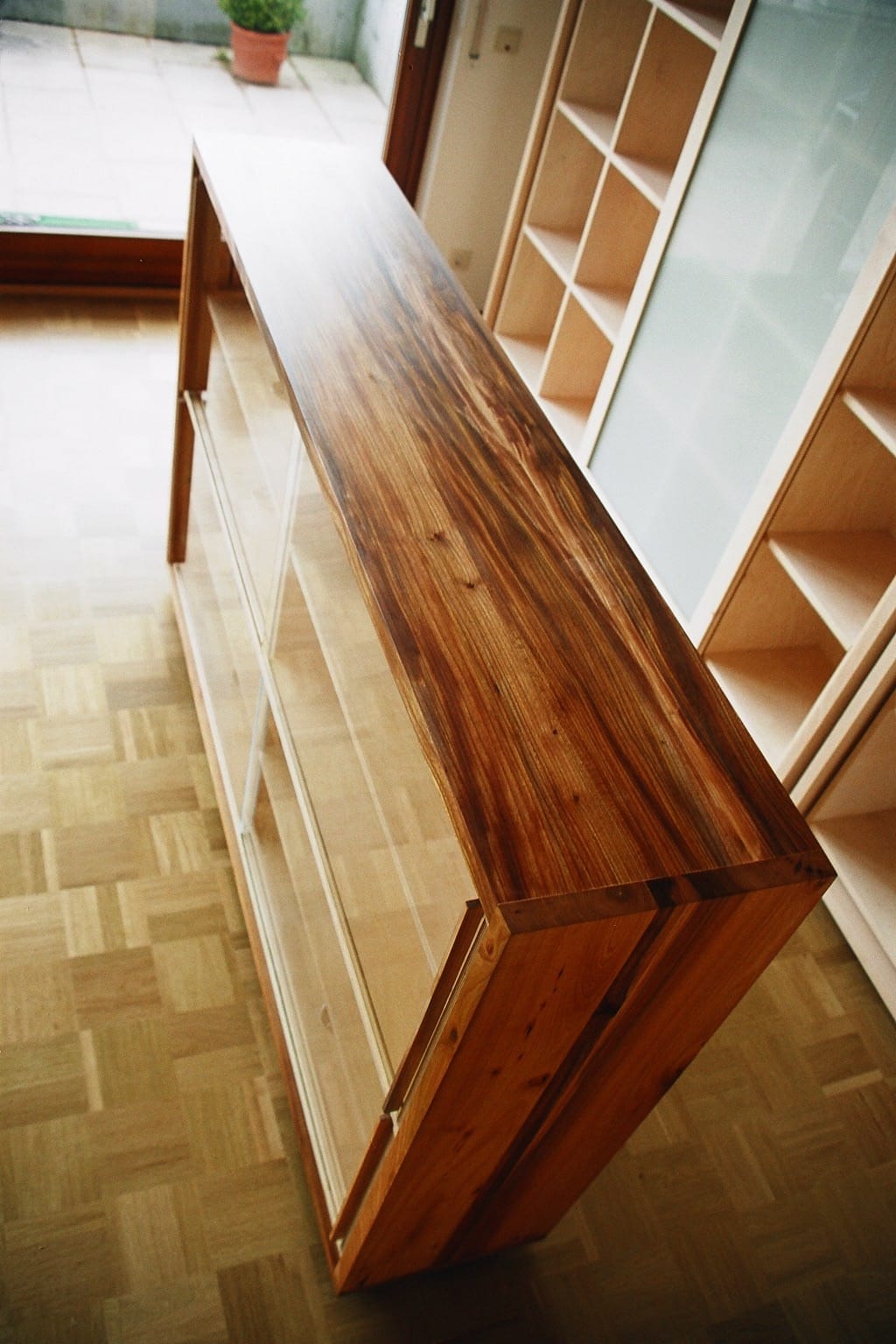 Eine Holzvitrine mit Glastüren steht in einem Raum mit Parkettboden, neben großen Fenstern und leeren Regalen.